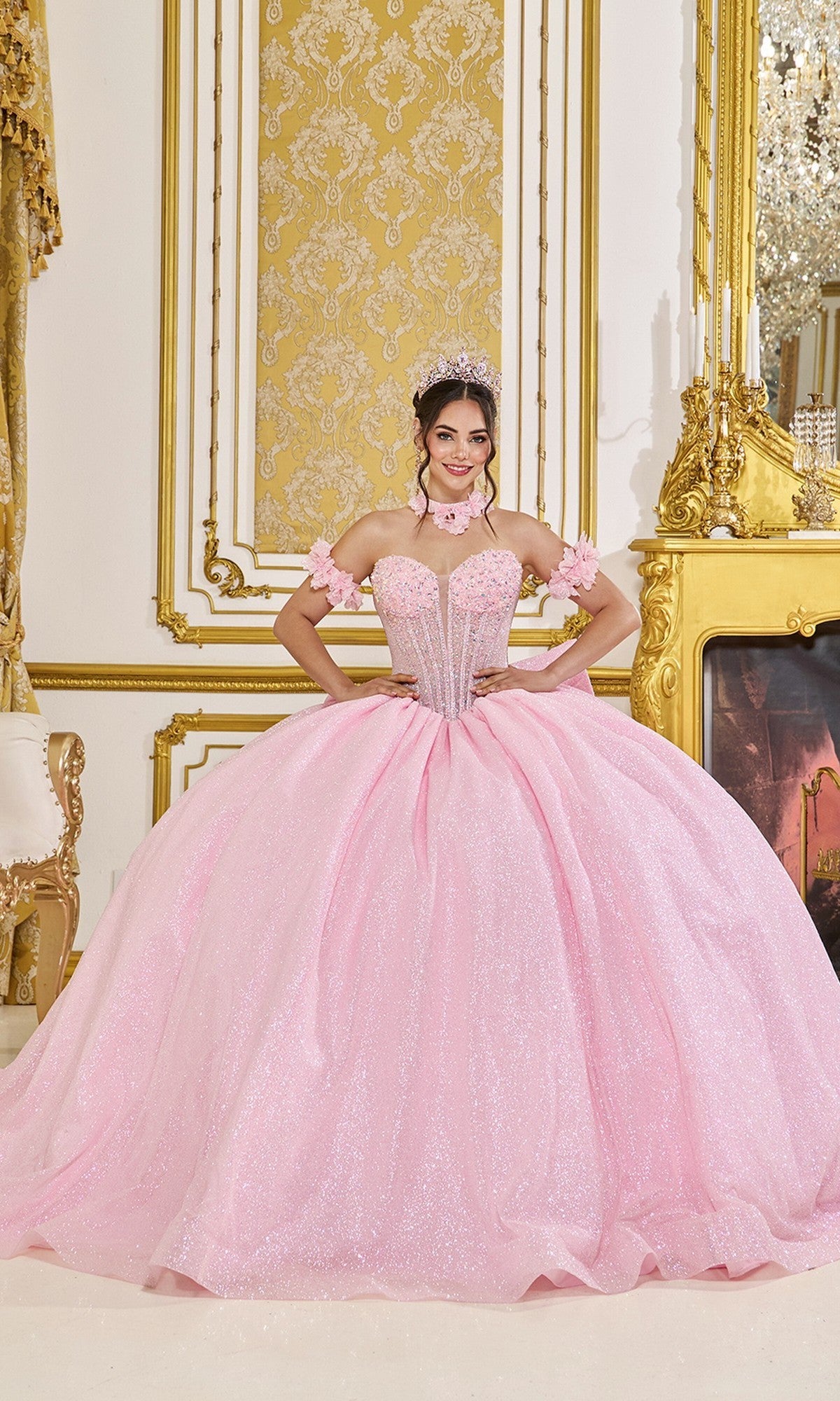 A young woman wears the Quinceanera Dress 5115 By Dancing Queen in pink, smiling at the camera as she stands in an ornate room with gold decor and a fireplace.
